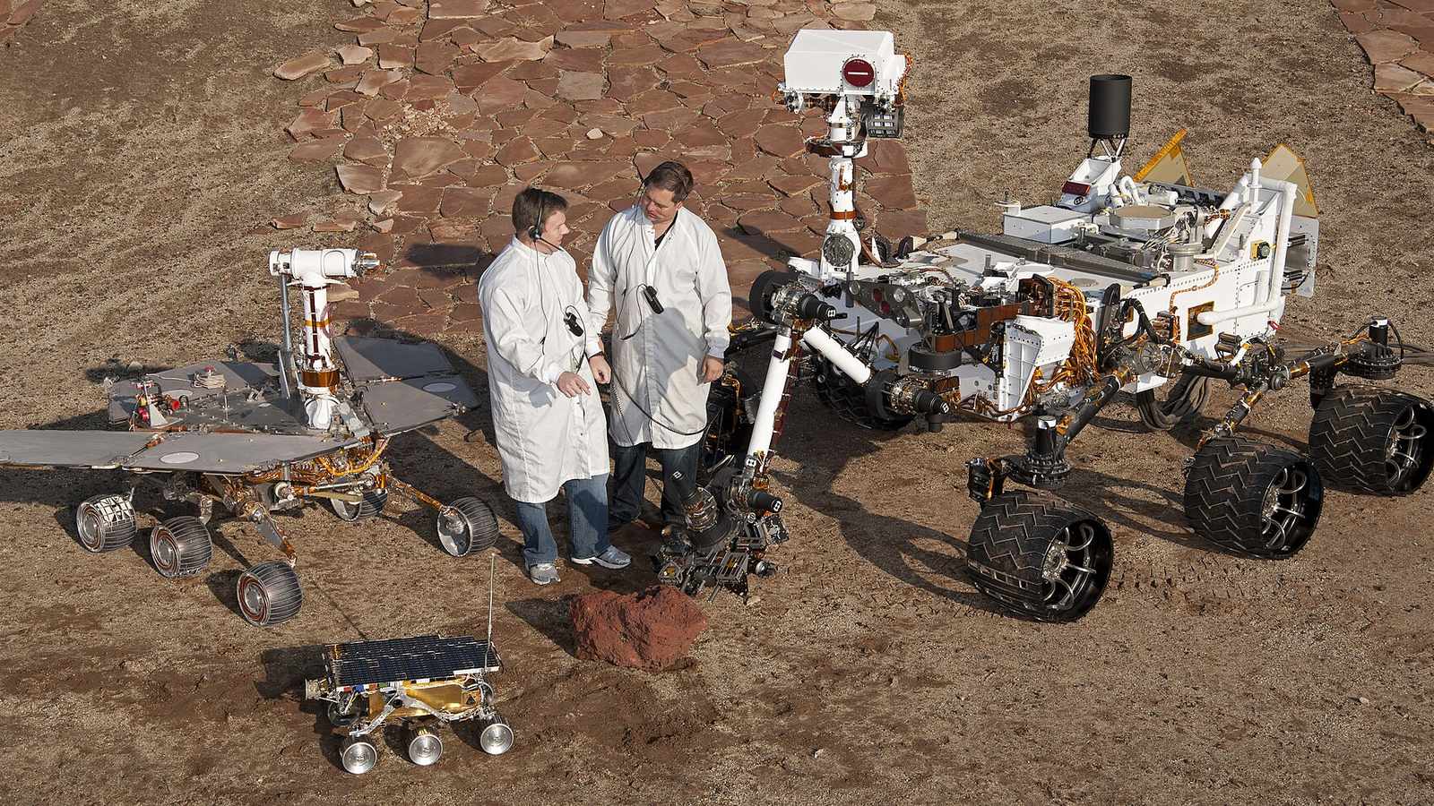 Two engineers in white coats stand in a rocky test yard among three NASA rovers of different sizes, a tiny Sojourner spare in front, a Spirit and Opportunity test rover on the left and a large Curiosity test rover on the right
